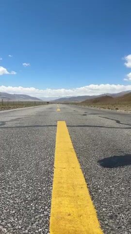 A low-angle shot on a long, straight asphalt road shows a single continuous yellow line down its center. The sky is clear blue with scattered white clouds, and distant mountains frame the horizon under a bright sun.

A person, seen from the waist down, walks directly away from the camera, straddling the yellow line. They wear dark, loose-fitting pants and white sneakers. The camera captures their footsteps, focused on their shoes.

The shot then transitions to a full-body view of the person, walking away from the camera along the yellow line. They are wearing a colorful, patterned long-sleeved top and the same dark, loose pants.

Suddenly, the person jumps high into the air, arms raised in a V-shape, pointing upwards. They are silhouetted against the bright sky, suspended mid-air above the yellow line, with the vast, open road and mountainous landscape stretching out behind them. The scene suggests a remote, possibly high-altitude location in Mainpu township, China, given the context. The weather is clear and sunny.