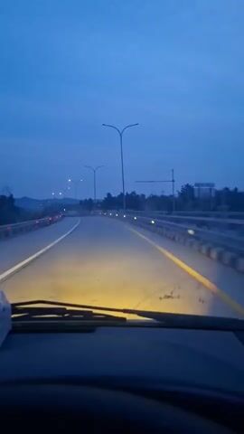 The video is a first-person perspective shot from inside a vehicle, driving on a road at dusk or night. The headlights illuminate the road ahead, casting a yellow glow. Streetlights line the road, and the sky is a deep blue. The road appears to be a highway or a well-maintained road with clear lane markings. The surrounding environment is dark, with hints of trees and possibly hills in the distance. The dashboard and windshield wipers are visible in the foreground.