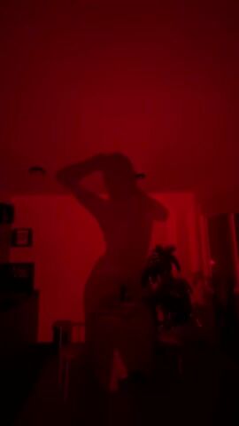 A person is dancing on a table in a room lit with red light. The person is silhouetted against a white wall. A table, a chair, and a plant are visible in the dimly lit foreground. There are pictures and a TV on the wall in the background. The person raises their right arm in the air. The room is dark save for the red lighting. Upbeat music plays.