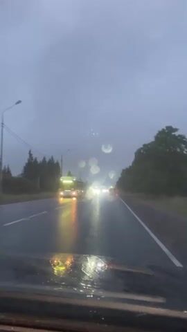 A dashcam view of a wet road at dusk with cars driving in the rain. The road is dark and reflects the headlights of oncoming vehicles. Trees line the right side of the road, and a few streetlights are visible in the distance. The sky is overcast and gray. A bus is visible in the distance on the left side of the road.