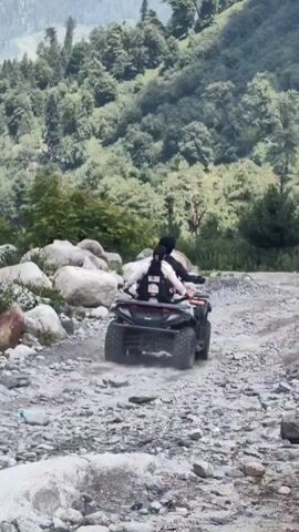 A quad bike is being ridden on a rocky path on a mountain slope. The quad bike is black and has two people on it. The person in the front is driving and the person in the back is wearing a backpack. The quad bike is moving away from the camera and up the slope. The mountain slope is covered in trees and rocks. The sky is cloudy.
