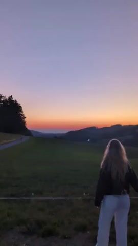 A woman with blonde hair stands in a grassy field, facing away from the camera. The sky behind her is a gradient of purple and orange, indicating either sunrise or sunset. In the distance, rolling hills are visible. The woman is wearing a dark jacket and white pants. She moves her arms, first outwards and then backwards, as if dancing or posing for a photo. There's a wire fence in front of her. To the left, a path and trees are visible.