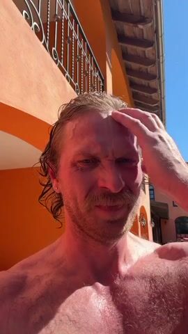 A shirtless man with red skin, a mustache, and wet blond hair is standing outside, facing the camera. He touches his hair, smiles, and looks around. The background features a tiled patio, an orange building with archways, and hanging lanterns. A small table is visible in the background, and a glimpse of a white dog appears briefly. The scene is brightly lit by sunlight.