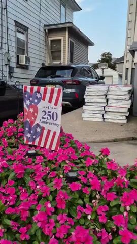 The video pans across the front of a residential house decorated for a celebration, likely the 250th anniversary. In the foreground, a bed of vibrant pink flowers is visible. To the left, a flag with red and white stripes and blue hearts with white stars is displayed, along with text that reads "th 250 Anniversary". Behind the flag, a black SUV is parked. A stack of white boxes is visible next to the SUV. The video then moves to the right, showing a set of brick stairs leading up to a porch. The porch and stairs are adorned with American flags and potted plants filled with red and white flowers. The house itself is a light-colored siding with white trim. The scene appears to be set in a suburban environment with other houses visible in the background. The lighting suggests it is either early morning or late afternoon. There are no people visible in the video.