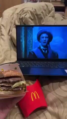 The video shows a person watching a movie on a laptop while eating a McDonald's meal in bed. The laptop screen displays a scene from the movie "Harry Potter," with close-up shots of Harry Potter and Professor Quirrell. The person's hand holds a Big Mac burger, with a bite taken out, near a container of french fries, both from McDonald's. The bed has a beige comforter.
