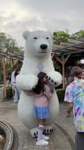 A girl in a pink jacket and blue skirt hugs a person in a white polar bear costume. The girl has long dark hair and is smiling. She is wearing white socks with black hearts and white sneakers. The polar bear costume is large and fluffy with black eyes and a black nose. The background shows a park-like setting with trees, wooden structures, and other people. There is a stone path in the foreground. Another child, a boy in a red shirt, is standing to the right of the girl and the bear. The sky is visible in the upper part of the frame.