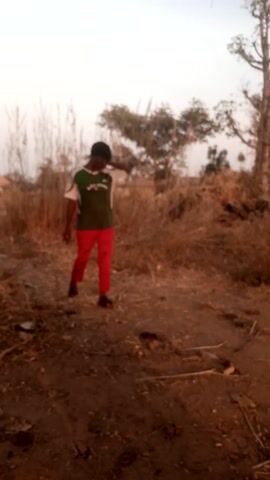 A woman is walking toward the camera on a dirt path. She is wearing a dark green t-shirt with white trim and the word "COYZUDA" across the front. She also has on bright red pants, black sandals, and a black head covering. The background consists of dry brush, a few trees, and a light sky. The shot is taken from a low angle, with some tree branches visible at the very top.