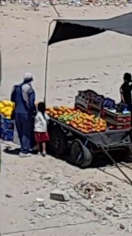 The video shows an outdoor scene with a fruit stand. A cart filled with oranges and other produce is set up under a black awning. Several people are present, including a person in a blue robe and head covering, and another person in a black shirt. A bicycle is also visible in the background. The ground is dusty and appears to be a street or market area. The overall impression is one of a busy marketplace on a sunny day.