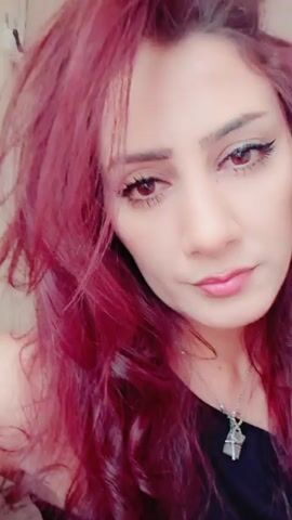 The video displays a close-up of a woman's face. Her hair is long and dyed a vibrant reddish-pink color. She has striking brown eyes with noticeable eyeliner and mascara. Her lips are painted a soft pink. She is wearing a black top, and a silver necklace with a pendant is visible. The background is indistinct, suggesting it's likely an indoor setting with diffused lighting. The camera angle is slightly angled, focusing on the woman's upper body and face. The video appears to be a selfie or a short, personal video clip.
