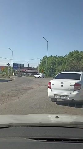 A white car is parked on the right side of a dirt road in a sunny outdoor environment. The car is facing away from the camera. In the distance, there are several other cars, a gas station, trees, and streetlights. The sky is clear and blue.