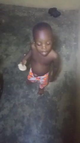 A young child is standing in a corner of a room. The child has dark skin and is wearing patterned shorts. The child is holding a piece of white material in their left hand. The child looks down and then up at the camera. A pair of dark shoes sits in the background of the room. The floor is gray.