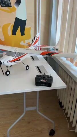 A remote-controlled airplane and its controller sit on a white table in front of a window. A woman laughs at the camera as she records a selfie.