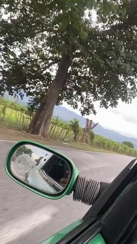 The video is a first-person perspective from inside a moving green vehicle, looking out through the driver's side window and side mirror. The side mirror reflects the road ahead and the interior of the car, including a person's hand holding a phone. The car is on a paved road that curves to the right. To the left of the road, there are fields and a fence, with a leafless, dead tree standing in the distance. Beyond the fields, there are rolling hills and mountains. The sky is overcast with dramatic clouds. As the video progresses, a truck passes the camera vehicle, followed shortly by a motorcycle with two riders. Power lines and utility poles are visible along the side of the road. The vegetation appears lush and green, suggesting a rural or natural environment. The overall impression is of a scenic drive through a somewhat rugged landscape under a cloudy sky.