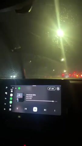 The video captures an interior view from a car at night, looking out through the windshield. The dominant feature in the foreground is an infotainment screen displaying a music player interface. The screen shows the song "American Girls" by "Harry Styles," with a progress of 3:13 out of 3:33. The play/pause button indicates the music is currently paused. Other elements on the screen include a digital clock displaying "8:11," navigation and phone icons on the left sidebar, and a "Jam" button at the top right of the music interface.

Through the car's windshield, the outside environment is dark and appears to be rainy or foggy, as water droplets are visible on the glass, and lights are diffused and hazy. Bright, circular streetlights illuminate the wet surface, casting a strong glare on the windshield. In the distance, several red taillights from other vehicles are visible, suggesting traffic or a populated road. The overall scene conveys a nocturnal car journey in challenging weather conditions, with music being played inside the vehicle.