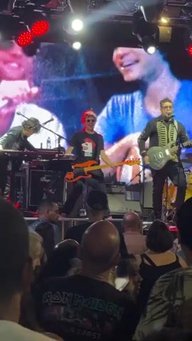 A band performs on a stage in front of a large screen displaying a black and white image of people. The screen is illuminated by stage lights, some of which are red. The band members are visible: a person on the far left appears to be playing a keyboard, a person in the center is playing a bass guitar and singing into a microphone, and a person on the right is playing an electric guitar and singing into a microphone. The audience is visible in the foreground, with their backs to the camera, facing the stage. The overall atmosphere is that of a live concert.