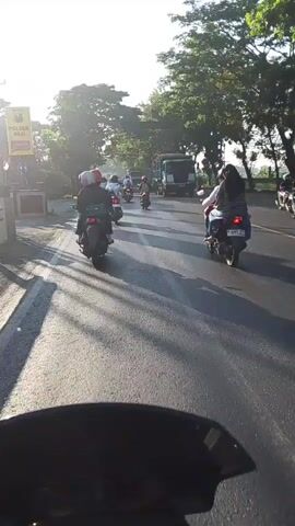 The video shows a POV shot from a motorcycle on a sunny day, riding on a paved road. Several other motorcycles are visible, some with passengers, moving in the same direction. A large truck is in the distance on the right side of the road. Trees line both sides of the road, with their shadows stretching across the asphalt. The sun is low in the sky, casting a warm glow and long shadows. The overall impression is of a busy road with moderate traffic in a rural or suburban setting.