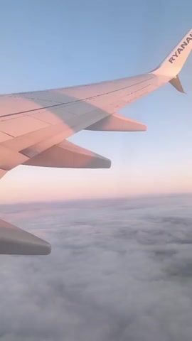 The video shows a view from an airplane window looking out at the clouds during sunrise or sunset. The wing of the airplane, branded with "RYANAIR", is visible in the foreground, casting a pink hue onto the clouds below. The sky is a clear blue, transitioning into soft pink and orange tones near the horizon. The clouds appear to be a thick, uniform layer stretching as far as the eye can see.