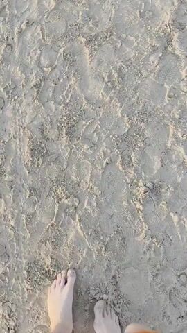The video shows a first-person perspective of someone walking on a sandy surface. The sand is textured with numerous footprints, some appearing to be from shoes with a distinct tread pattern and others from bare feet. The camera pans slightly downwards, showing the walker's bare feet and legs, wearing orange shorts. The lighting suggests it is daytime, possibly late afternoon, with soft shadows. The environment appears to be a beach or a similar sandy area.