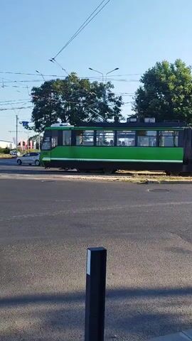 The video captures a sunny day in Bucharest, Romania, likely during late morning or early afternoon, given the clear blue sky and bright sunlight casting distinct shadows. The scene is an urban intersection with tram tracks prominently featured. A green articulated tram is the central object, moving across the frame from left to right. Its windows are visible, showing glimpses of passengers inside.

Several vehicles are present, including a white van and a dark-colored car, suggesting moderate traffic. Overhead, a complex network of power lines for the tram is evident, along with streetlights and traffic signals. In the background, trees line the road, adding greenery to the urban landscape. A McDonald's sign is visible on a distant building, providing a recognizable location cue.

A person is seen walking into the frame from the right in the foreground, partially obscuring the view of the tram and intersection. They are wearing a light blue shirt and carrying a dark bag. Their presence suggests that this is a pedestrian area or a crosswalk. The overall atmosphere appears to be one of ordinary urban life in progress, with the focus on the movement of public transportation and the daily activities of the city. No text is clearly legible within the video, other than the McDonald's logo.