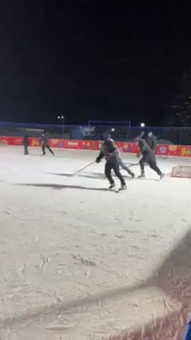 The video shows an outdoor ice rink at night with several people playing hockey. They are dressed in winter clothing and using hockey sticks to maneuver a puck on the ice. The rink is illuminated by bright overhead lights, and there's a decorative border around the perimeter of the ice surface. The ice appears well-maintained, and the players are actively engaged in the game.