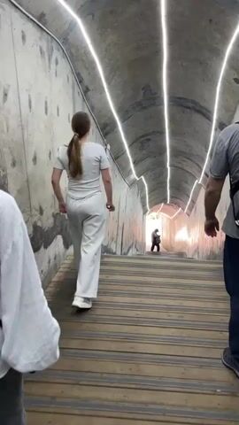 A low-angle shot shows a person walking down a long, lighted tunnel staircase. The tunnel has a gray, concrete ceiling that curves over the stairs, with long, bright white lights attached to the ceiling. The stairs are made of wooden planks. A young woman wearing a gray t-shirt and loose gray pants is walking down the stairs in front of the camera. Her light brown hair is pulled back in a ponytail. To the right of her is an adult male wearing a gray shirt and blue pants. To the left, another person is partially visible in a white striped shirt. Far down the stairs, another person is visible, dressed in dark clothing. The background light at the end of the tunnel is very bright, making it hard to see.