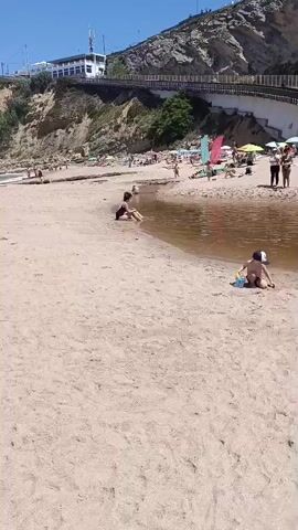 The video shows a sandy beach on a sunny day. A small river or stream runs into the ocean. Several people are on the beach, some sunbathing and others playing in the water. Children are seen playing in the sand and in the shallow water. In the background, there are cliffs with a wooden walkway or bridge built along them. Some umbrellas and beach chairs are scattered on the sand.
