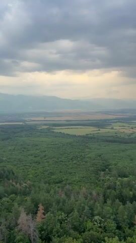 This is a vertical video clip that appears to be a selfie video taken from a high vantage point. The initial frames showcase a panoramic landscape of rolling green hills, a forested area, and a distant town nestled in a valley. The sky is dramatic with heavy, grey clouds interspersed with patches of warm, golden light, suggesting either sunrise or sunset.

As the video progresses, the perspective shifts to include the person filming. A young man with curly dark hair and a black tank top is prominently featured, smiling and looking towards the camera. The background behind him is a blur of green foliage and a dirt path winding through the landscape, consistent with the earlier scenic views.

The video then cuts to a closer view of the structure the person is standing on, revealing metal beams and a platform, indicating they are atop a tower or a similar elevated structure. The final frames briefly return to the sky and distant landscape, emphasizing the height and the expansive view.

The overall mood is one of adventure and appreciation for nature, with the dramatic sky adding a sense of awe. The time of day is likely early morning or late evening due to the lighting. The location context provided is Turiya, Bulgaria, which aligns with the picturesque, rural scenery depicted. There is no visible text in the video.