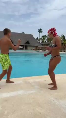 A woman and a boy are mimicking boxing moves next to a swimming pool. The boy is on the left wearing green shorts with yellow designs, and he is shirtless. The woman is on the right wearing a leopard-print bikini, goggles, and a red and white head covering. They are both barefoot. The swimming pool is turquoise, and in the background, there is a building with a thatched roof and palm trees. The sky is cloudy. They are facing each other and bobbing up and down, with their fists up as if boxing.