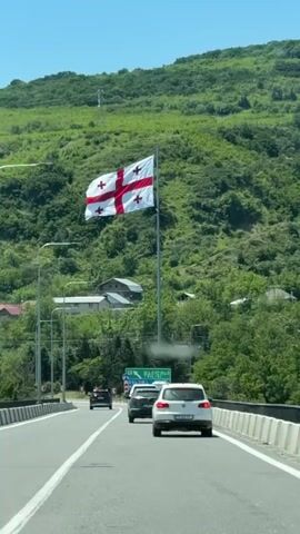A person is driving on a highway on a sunny day. A large Georgian flag is flying from a tall flagpole on the right side of the road, with green mountains in the background. Other cars are also visible on the highway.