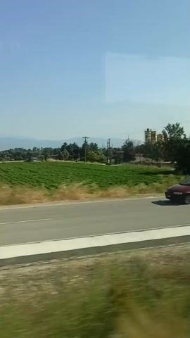 The video shows a view from a moving vehicle, likely a car or bus, looking out a window at a landscape. The scene is dominated by a clear blue sky and a green field, possibly agricultural. A road runs horizontally across the middle of the frame, separating the foreground from the background. Beyond the green field, there are trees and some buildings in the distance, including what appears to be a tall, yellow structure. The foreground shows blurred, dry vegetation, indicating the motion of the vehicle. The overall impression is of a rural or semi-rural setting on a sunny day.