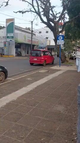A street scene at a Petrobras gas station. A red hatchback car is parked at a gas pump. A woman in a white top and light-colored pants walks along the sidewalk, facing the camera. Another car, a dark-colored hatchback, is parked on the street to the left. A sign with the letter "E" is visible on a blue pole. Trees with foliage are present in the background. The ground is paved with square tiles.
