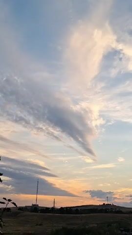 A time-lapse video shows a sky filled with wispy clouds and a hint of sunset colors. The bottom of the frame reveals a dark, silhouetted landscape with what appear to be distant buildings and utility poles. The clouds are dynamic, shifting and changing shape as the video progresses, creating an ethereal and atmospheric scene.