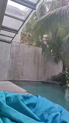 A video begins showing a blue beanbag chair in the foreground. To the right is a swimming pool with clear blue water and a wooden deck. Palm trees and other tropical foliage are visible in the background. The scene is outdoors, likely in a tropical climate. The camera is positioned low, as if the viewer is sitting in the beanbag chair. The footage is shaky and appears to be handheld.