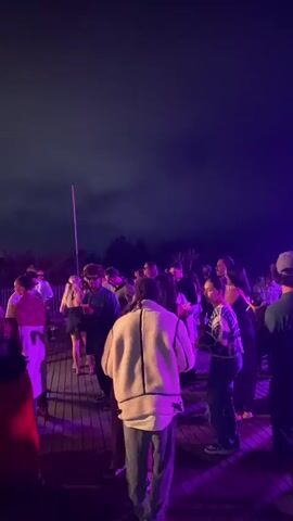 The video shows a group of people dancing at an outdoor event at night. The scene is illuminated by purple and pink lights, suggesting a party or concert atmosphere. The sky is dark, and the ground appears to be a wooden deck or platform. People are dressed casually, and some are interacting with each other. There are speakers visible, indicating that music is playing.