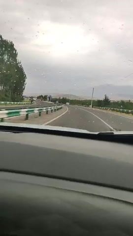 This media file captures a view from inside a vehicle driving on a highway. The scene is set in Shahrinav, Tajikistan, under a cloudy, overcast sky, suggesting it might be morning or late afternoon, with the weather appearing to be rainy or recently rained on, as evidenced by water droplets on the windshield.

The highway itself is asphalt with clear white lane markings. On the left side of the road, there's a green metal guardrail and a line of trees. Further down the road on the left, a few cars and trucks are visible, as well as what appears to be a billboard with a large image on it, possibly depicting a person. The right side of the highway also features a green guardrail and sparse trees. In the distance, rolling hills and mountains are faintly visible through the haze.

The primary activity is the act of driving along this highway, with the perspective of the camera indicating a forward-facing view from the driver's or passenger's seat. There are no specific people prominently featured as subjects, but other vehicles on the road indicate the presence of other travelers. The overall mood conveyed is one of travel and perhaps a bit of introspection due to the subdued weather conditions. The landscape suggests a semi-rural or exurban setting. No prominent text is legible on the billboard or any signage within the frame.