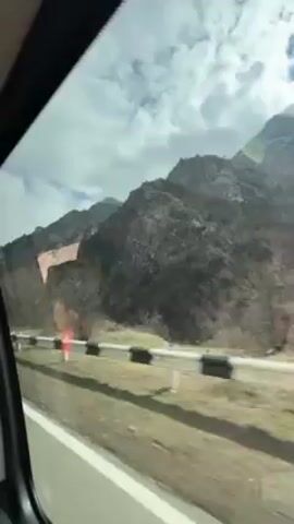 The video shows a view of mountains from a moving vehicle. The mountains are rocky and partially covered with vegetation. There is a road with a guardrail visible in the foreground. The sky is partly cloudy with patches of blue. The camera is positioned inside the vehicle, looking out through the side window. The video appears to be shot during the daytime. There is some glare on the window from the sun.