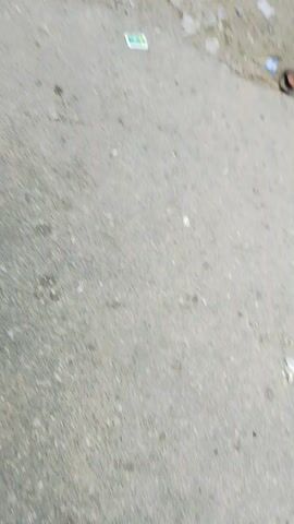 The video opens with a close-up of a person's hand holding a black plastic bag while standing on a gray, weathered surface that appears to be pavement. The person is wearing a blue garment and white shoes. The camera then pans to show the person's legs and feet, continuing to walk. The next shot shows a person in a white lace garment and head covering, standing next to a colorful auto-rickshaw. The auto-rickshaw has various decals and text on its sides, including "TVS". Another person is visible near the auto-rickshaw. The camera is tilted sideways, giving a skewed perspective of the scene. The environment appears to be an outdoor setting, possibly a street or roadside, with a glimpse of a sky in the background. There are other people and vehicles present, suggesting a public area. The footage then focuses on the person in the white lace garment, who is lying on the ground next to the auto-rickshaw. A hand wearing a bracelet is visible near the person in white. The person in the blue garment from the beginning of the video is also seen, holding another black plastic bag. The camera then pans to show a wider view of the auto-rickshaw, the person in white, and the person in blue. The scene appears to depict an incident or event, with the person lying on the ground.