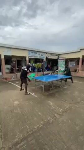 Two individuals are engaged in a game of table tennis outdoors. The setting appears to be a courtyard or open area surrounded by buildings. The table tennis table is blue. One person, wearing a cap, dark jacket, and shorts, is on the left side of the table. The other person, clad in a green outfit, stands on the right side. They are actively hitting the ball back and forth. Several people can be seen watching in the background. The weather appears overcast.