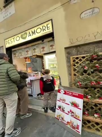 The video shows a street scene in Genoa, Italy. In the foreground, a small shop called "Pesto Per Amore Genova" is visible. A family, consisting of two adults and a child, are looking at the products inside the shop. The shop has a display of pestos, and a person wearing a red jacket is helping the customers. Next to the shop is a Christmas-themed decoration made of green foliage with red ornaments. The backdrop shows an old stone archway with a sign, a bright blue sky, and other buildings. People are walking under the archway. The video is shot from a slightly low angle, and the camera moves up and down.