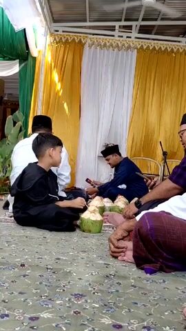 Here is a description of the video:

The video shows several males sitting on a carpeted floor, arranged in a circle around green coconuts. One of the men, dressed in a blue top, is reading something held in his hands, and the boy next to him, dressed in black, is focused on the coconuts. Other men are visible in the background, some in white clothing. The setting appears to be indoors, with yellow and white drapery forming a backdrop.