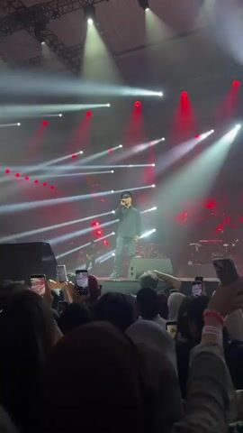 A singer is on stage in front of a large crowd, performing under bright stage lights. He is wearing a dark shirt, dark jeans, and a hat, and is holding a microphone. The crowd is holding up their phones to record him. There are other musicians on stage behind him, and large red and white light displays behind them. The venue appears to be an indoor arena.