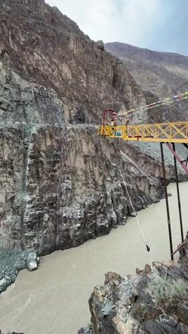 The video shows a person bungee jumping from a yellow platform suspended over a canyon. The canyon walls are steep and rocky. A muddy river flows at the bottom of the canyon. Prayer flags are attached to the platform. The jumper is secured by a rope and appears to be falling towards the river. The camera is positioned on a rocky outcrop overlooking the canyon, providing a view of the entire jump.