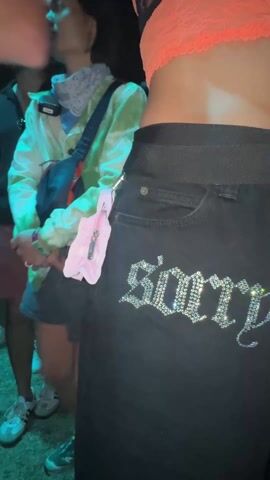 The video shows a person wearing black jeans with the words "sorry dad" bedazzled on the back. The person also has on a coral-colored top and a black belt. There are other people in the background, including one wearing a tie-dye jacket and another in a black shirt and red shorts. The scene appears to be in a crowded outdoor setting.