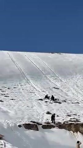 The video depicts a snow-covered mountainside under a clear blue sky. There are visible tracks in the snow, suggesting previous movement. Several people are visible on the mountainside. Some appear to be seated or kneeling, while others are standing near a rocky outcropping. The terrain is a mix of snow and exposed rock.