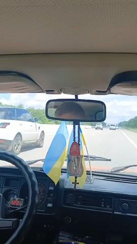 The video is filmed from the perspective of a passenger in a car, looking forward through the windshield. The dashboard of the car is visible, including the steering wheel and instrument cluster. Two Ukrainian flags are hanging from the rearview mirror, along with a religious icon and prayer beads. Outside the car, a highway is visible with other vehicles. The weather appears to be clear and sunny. The video is shot in a slightly shaky manner, suggesting it was filmed while the car was in motion.