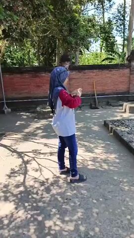 A person wearing a headscarf and a white and red shirt walks past an area with what appears to be gravestones. The area is paved with gravel and dirt, and there is a brick wall in the background. The person is holding a phone and appears to be taking a picture. Another person in a dark shirt stands near a lamppost in the background. The scene is outdoors, with trees and a blue sky visible. The gravestones are covered in gravel and appear to be very old. There are also piles of bricks and rubble scattered around the area.