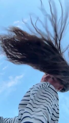 The video shows a person from a low angle, looking up at them against a clear blue sky with scattered white clouds. The person has long, dark hair that is being blown around by the wind, creating a dynamic and somewhat blurry effect. They are wearing a striped long-sleeved shirt and light-colored pants. The person is moving, with their head turning and their hair whipping around. The camera appears to be stationary, capturing the person's actions in relation to the sky.