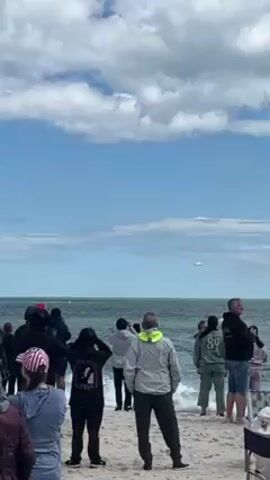 Here is a concise description of the video: 

The video shows a crowd of people standing on a beach, looking out towards the ocean. In the distance, an airplane is approaching. The sky is partly cloudy and the sea is calm.