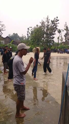 Here is a concise description of the video:

The video shows a group of people standing in a wet outdoor area. One person in the foreground, wearing a white t-shirt, shorts, and a cap, stands barefoot. Behind him, there are others wearing black shirts, some with masks. Further in the background, more people can be seen, along with trees and a building. The ground is wet and reflective, suggesting it has recently rained or is still raining. There's a blue-edged object in the upper right corner, possibly part of a structure.  The perspective is vertically oriented.