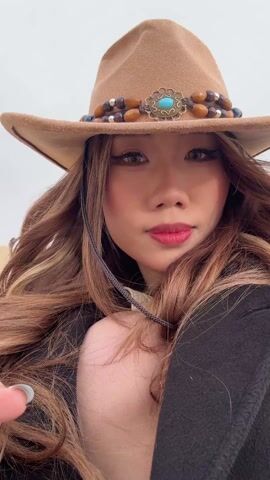 A young Asian woman with long brown hair, wearing a light brown cowboy hat decorated with beads and a turquoise embellishment, and a dark coat, poses for a selfie-style video. She has red lipstick and looks directly at the camera with a calm expression, sometimes smiling slightly.

The setting is a vast sand dune under an overcast sky. In one shot, the woman holds a smartphone, and the background reveals a panoramic view of the dune sloping down to an oasis-like area with buildings, water features, and some greenery. Other individuals can be seen in the distance on the sand dune. The scene suggests a desert environment, specifically the sand dunes near Dunhuang City, China, as indicated by the context.